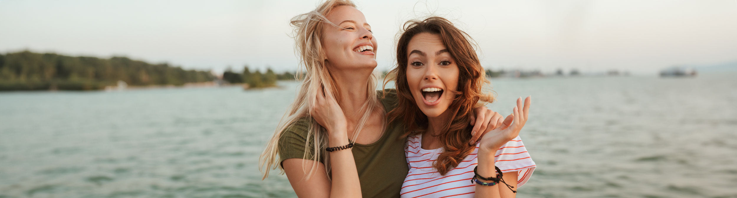 Zwei junge Frauen stehen dicht beieinander an einem Gewässer, lächeln und lachen. Eine trägt ein grünes Hemd, die andere ein weiß gestreiftes Hemd. Im Hintergrund sind Bäume und eine entfernte Uferlinie zu sehen.