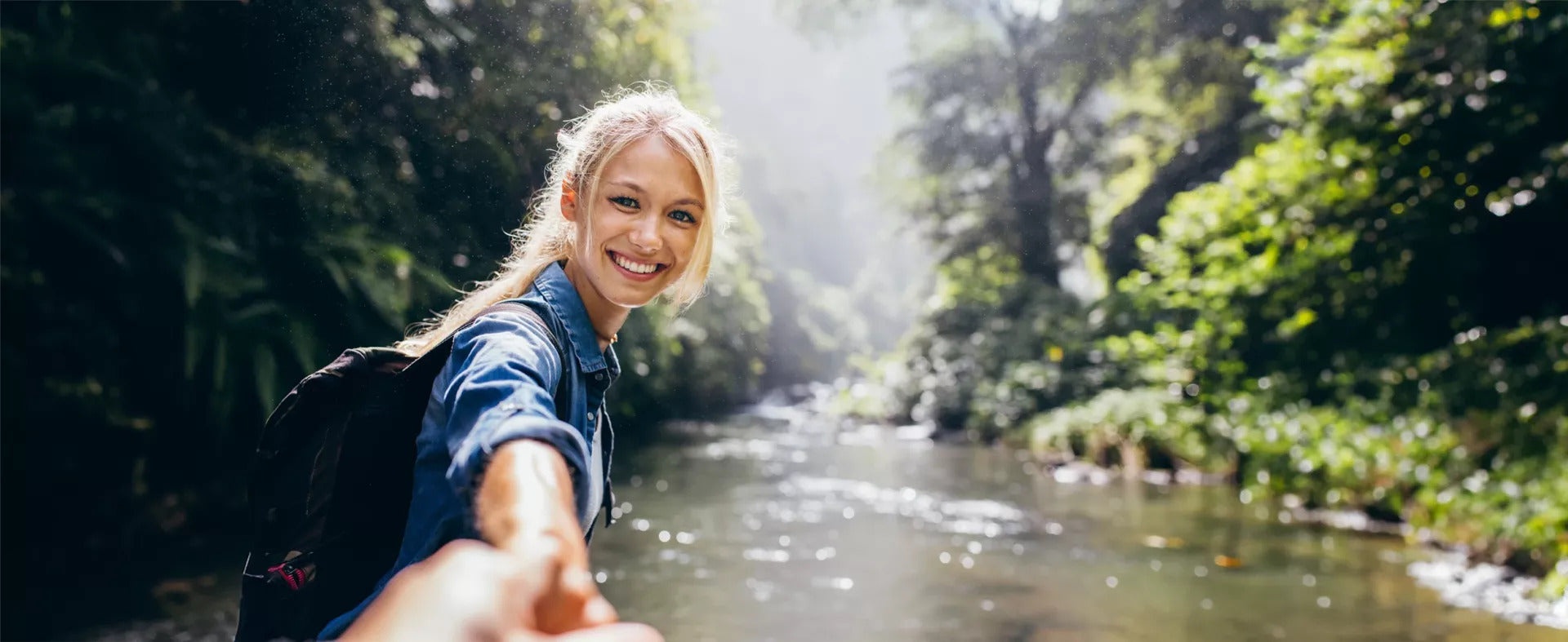 Eine lächelnde Frau mit Rucksack steht an einem Fluss in einem üppigen Wald. Sie streckt die Hand aus, hält jemanden an der Hand und lädt ihn ein, ihr zu folgen. Sonnenlicht fällt durch die Bäume und schafft eine helle, fröhliche Atmosphäre.