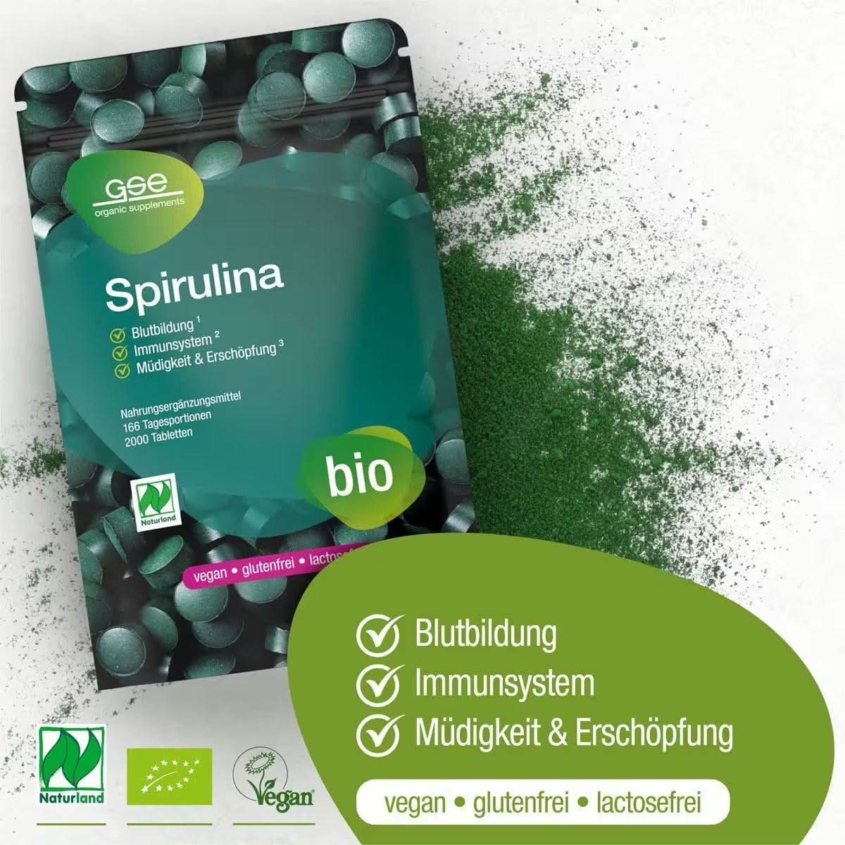 Eine Packung Naturland Bio Spirulina-Tabletten von gse.organic.supplements liegt auf einer weißen Fläche, daneben ist grünes Spirulina-Pulver verstreut. Auf der Verpackung werden Vorteile wie die Unterstützung der Blutbildung, der Immunität und die Verringerung von Müdigkeit erwähnt.